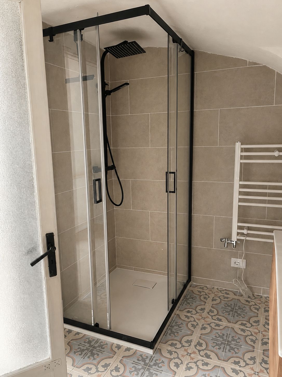 A glass-enclosed shower with a handheld showerhead, a white towel rack, and patterned floor tiles.