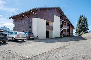 Ein zweistöckiges Holzhaus mit Balkon, geparkten Autos und Bergen im Hintergrund.