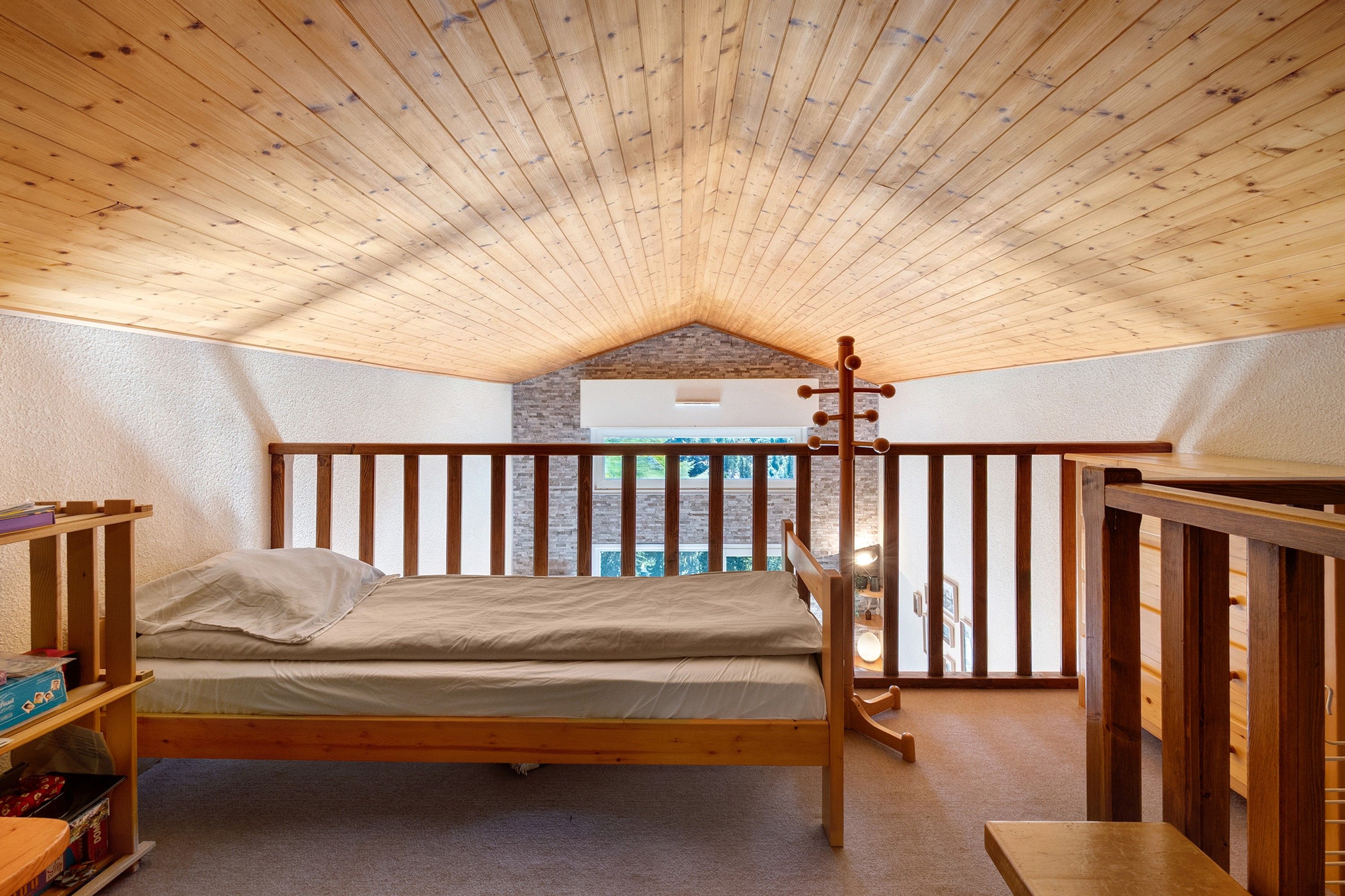 Une chambre avec un plafond en pente, un lit en bois, une petite table et une vue sur l’extérieur.