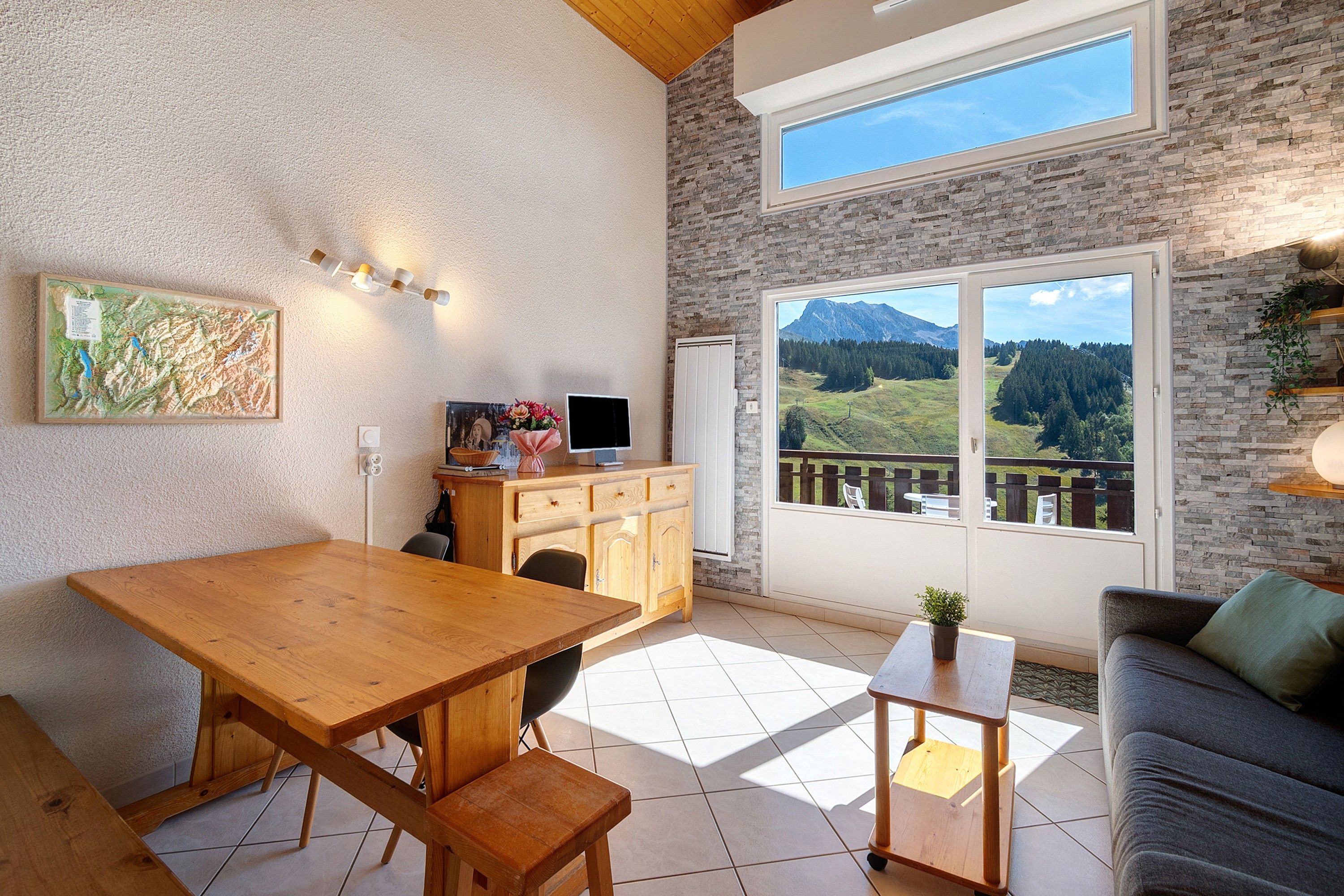 A cozy interior with a wooden dining table, a sofa, and a view of a mountainous landscape through large windows.