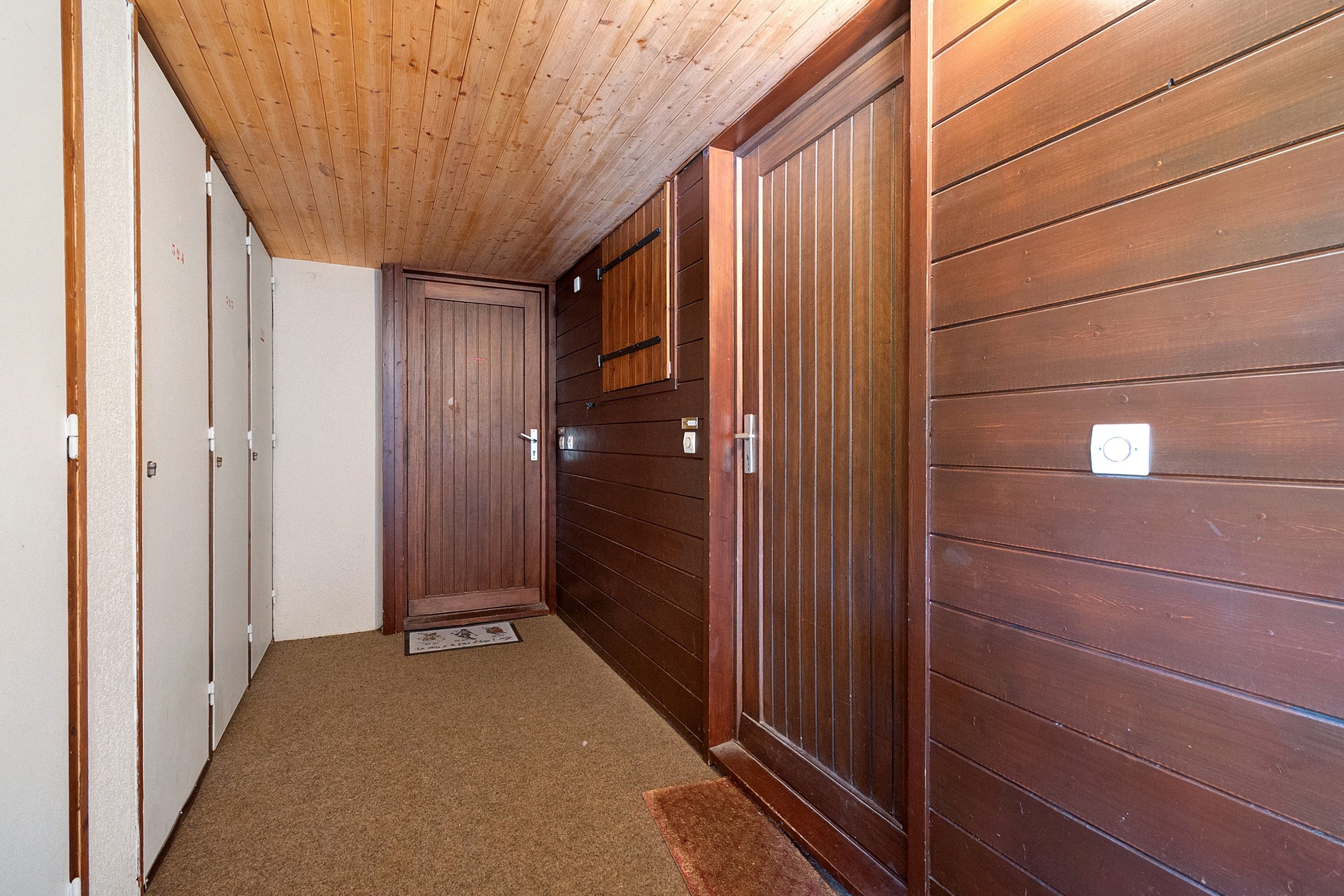 A narrow hallway with wooden walls and a wooden ceiling. There are doors on the left and a large wooden cabinet on the right.
