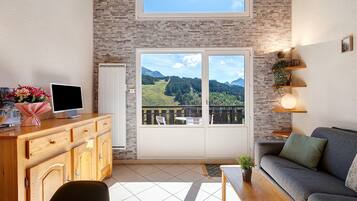 A living room with a wooden cabinet, a television, a sofa, a small table, and a balcony with a view of a mountainous landscape.