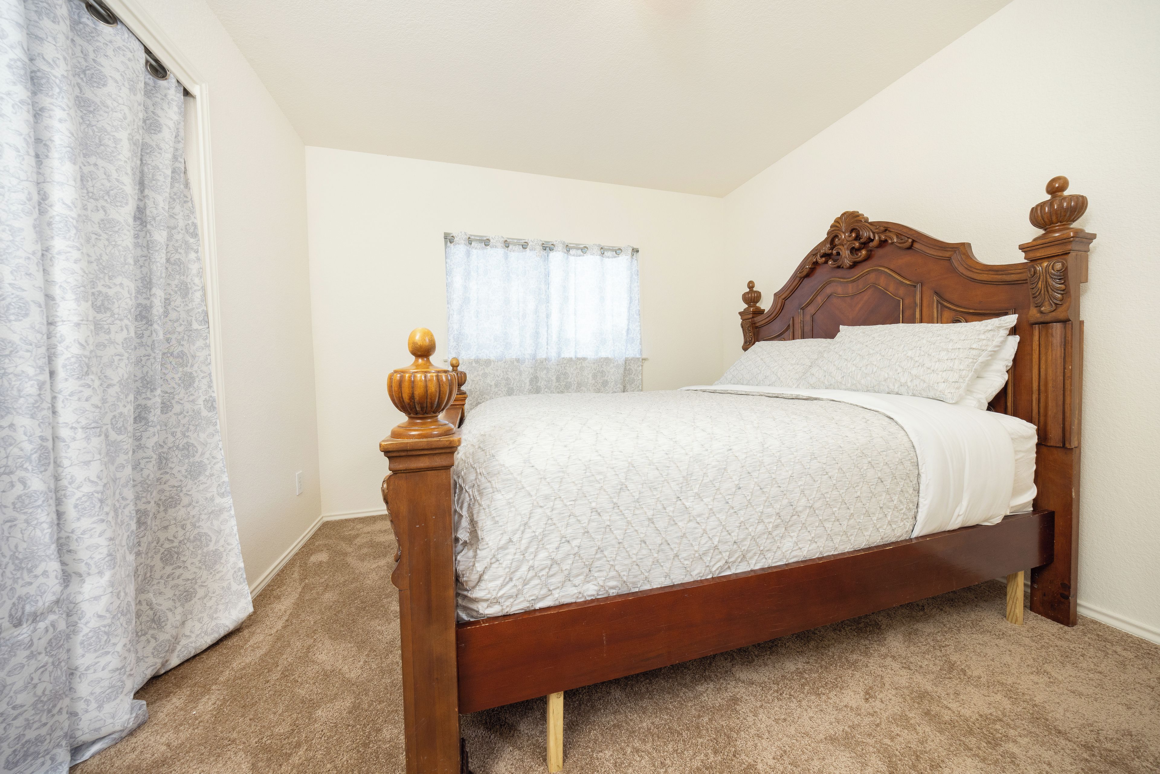 A bedroom with a wooden bed, a window with a curtain, and a carpeted floor.