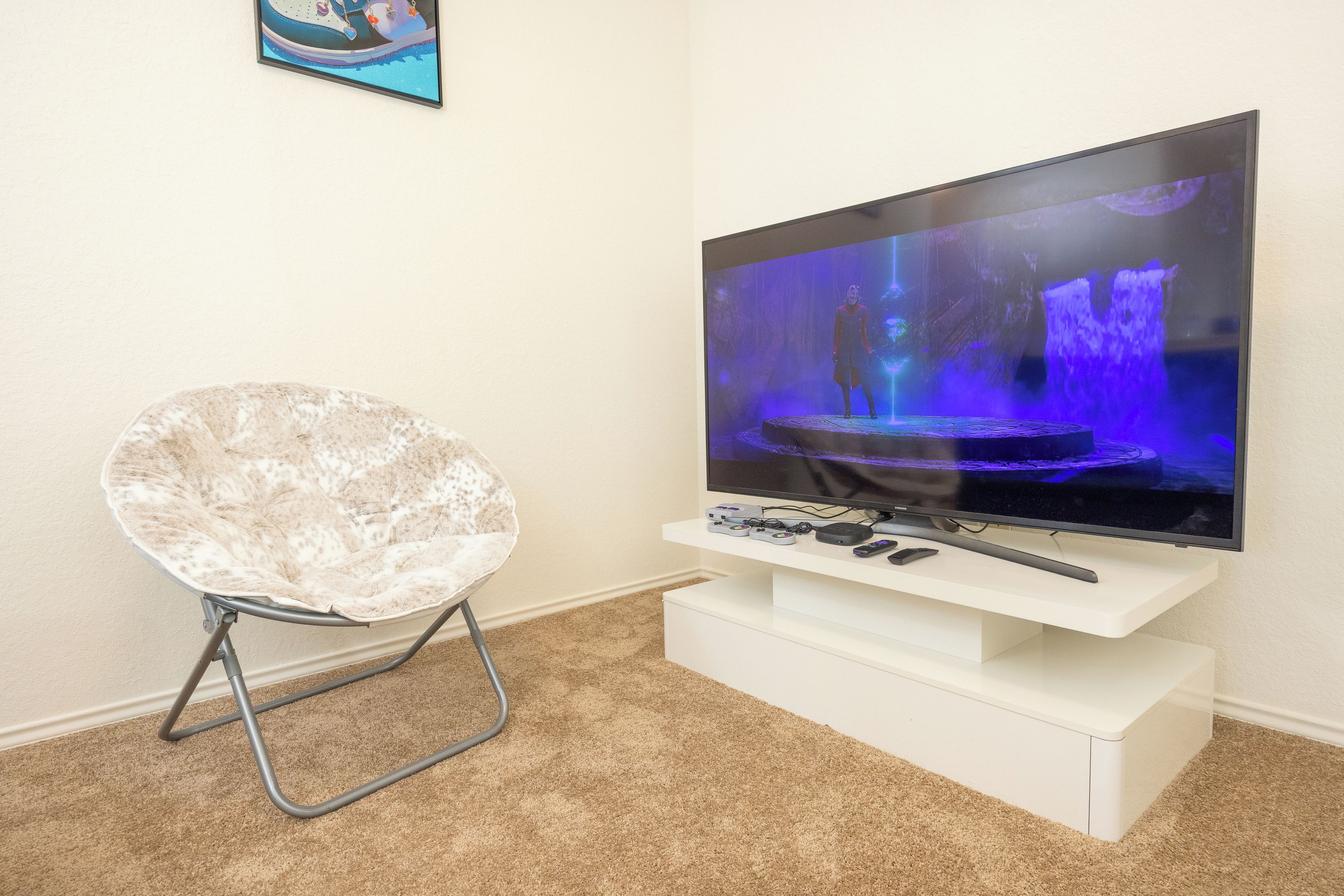Un téléviseur à écran plat posé sur un meuble blanc, avec une console de jeux et des télécommandes, une chaise blanche avec un siège moelleux et un sol recouvert d’une moquette.