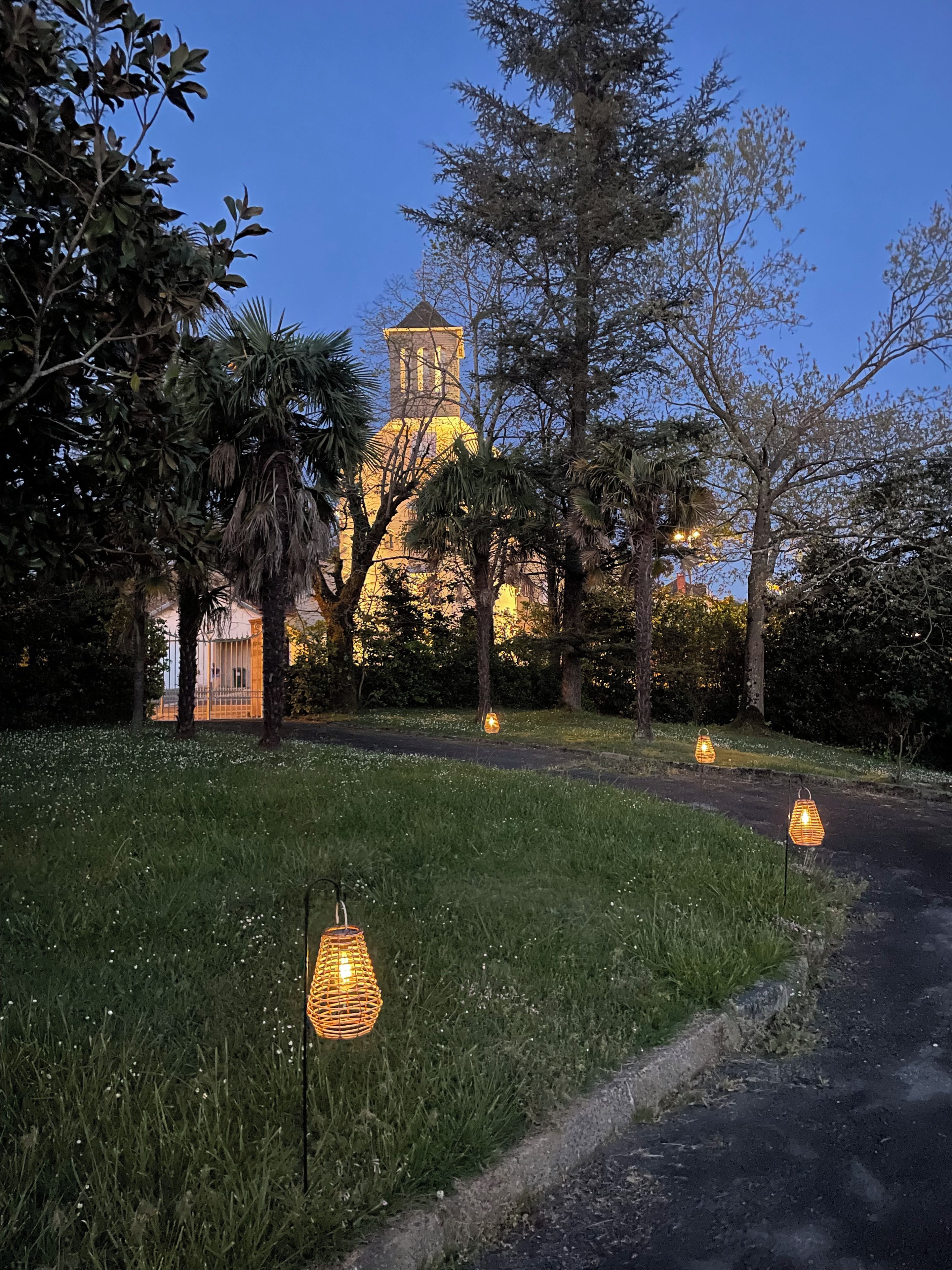 Un chemin menant à un bâtiment doté d’une tour, entouré d’arbres et illuminé par des lanternes suspendues.
