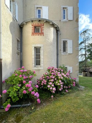A building with a curved facade, multiple windows with shutters, and a small door. There are lush pink flowers in front of the building.