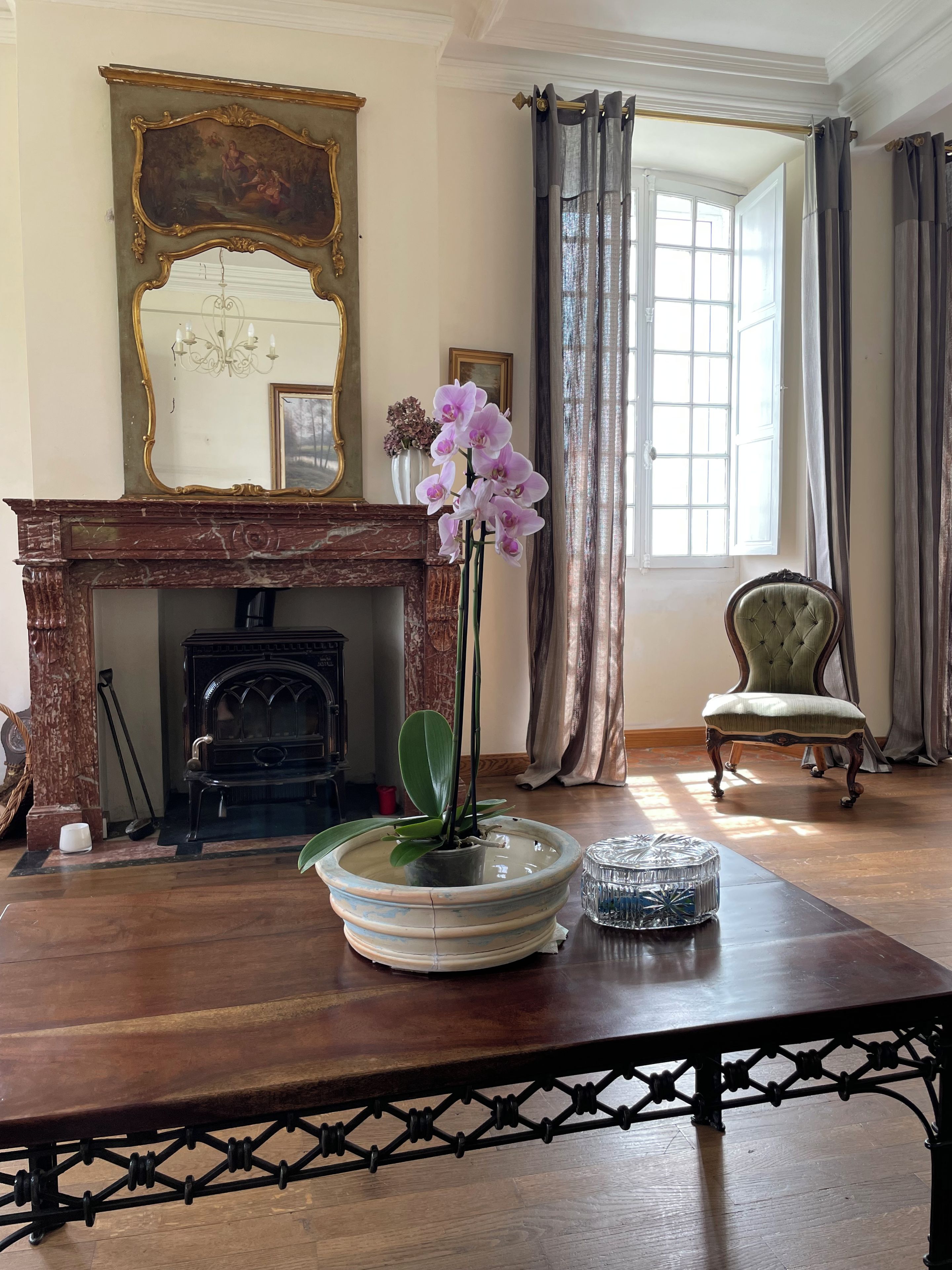 Een kamer met een open haard, een tafel met een bloemenvaas, een stoel en een raam met gordijnen.