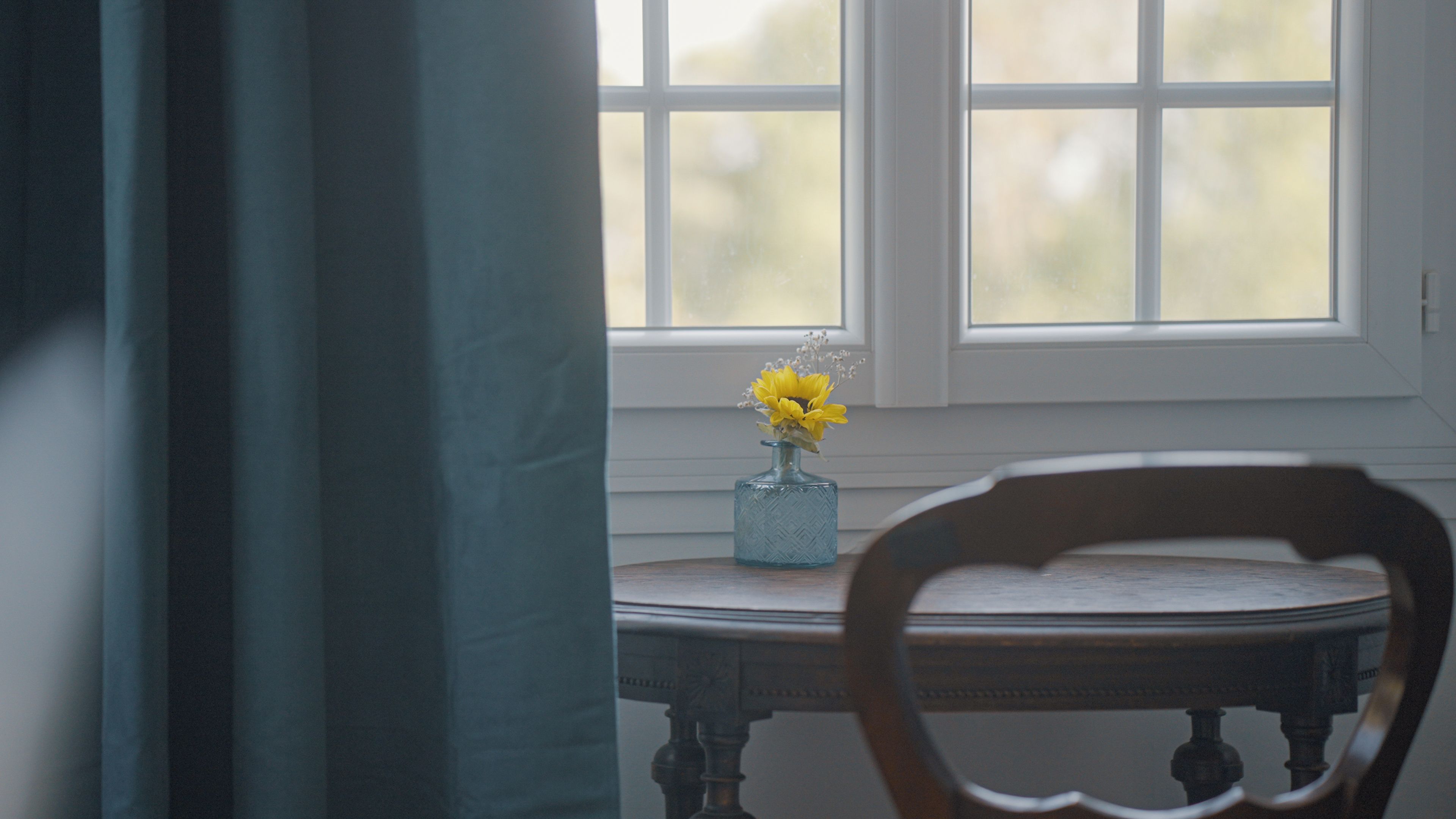 A room with a window, a table with a vase of flowers, and a chair.