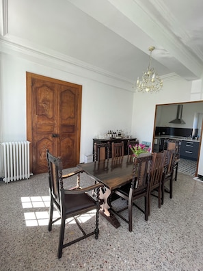 Een eetkamer met een grote houten tafel, zes stoelen, een kroonluchter en een keuken op de achtergrond.