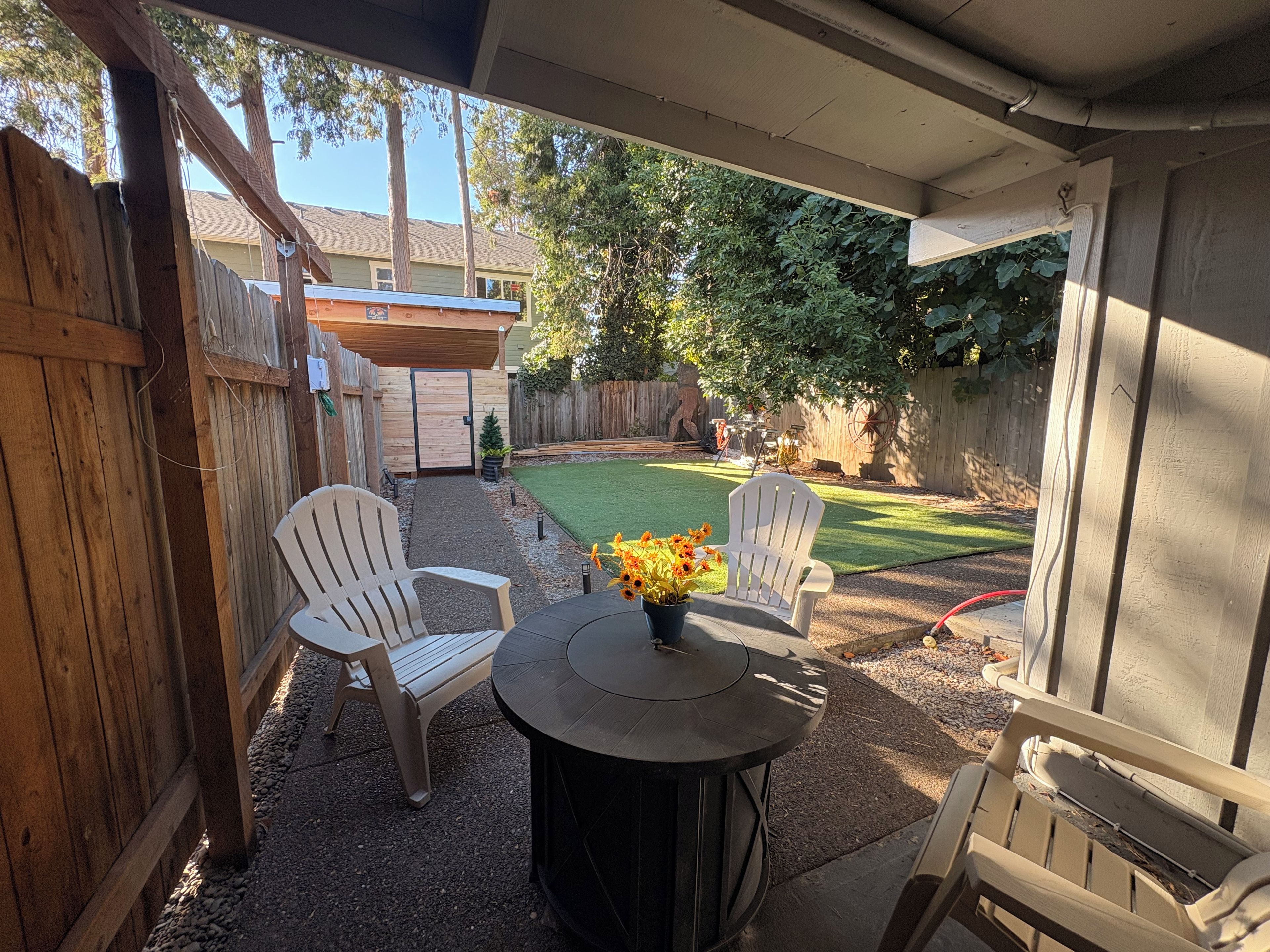 Un patio exterior con dos sillas blancas, una mesa redonda y un pequeño jardín con césped y árboles.