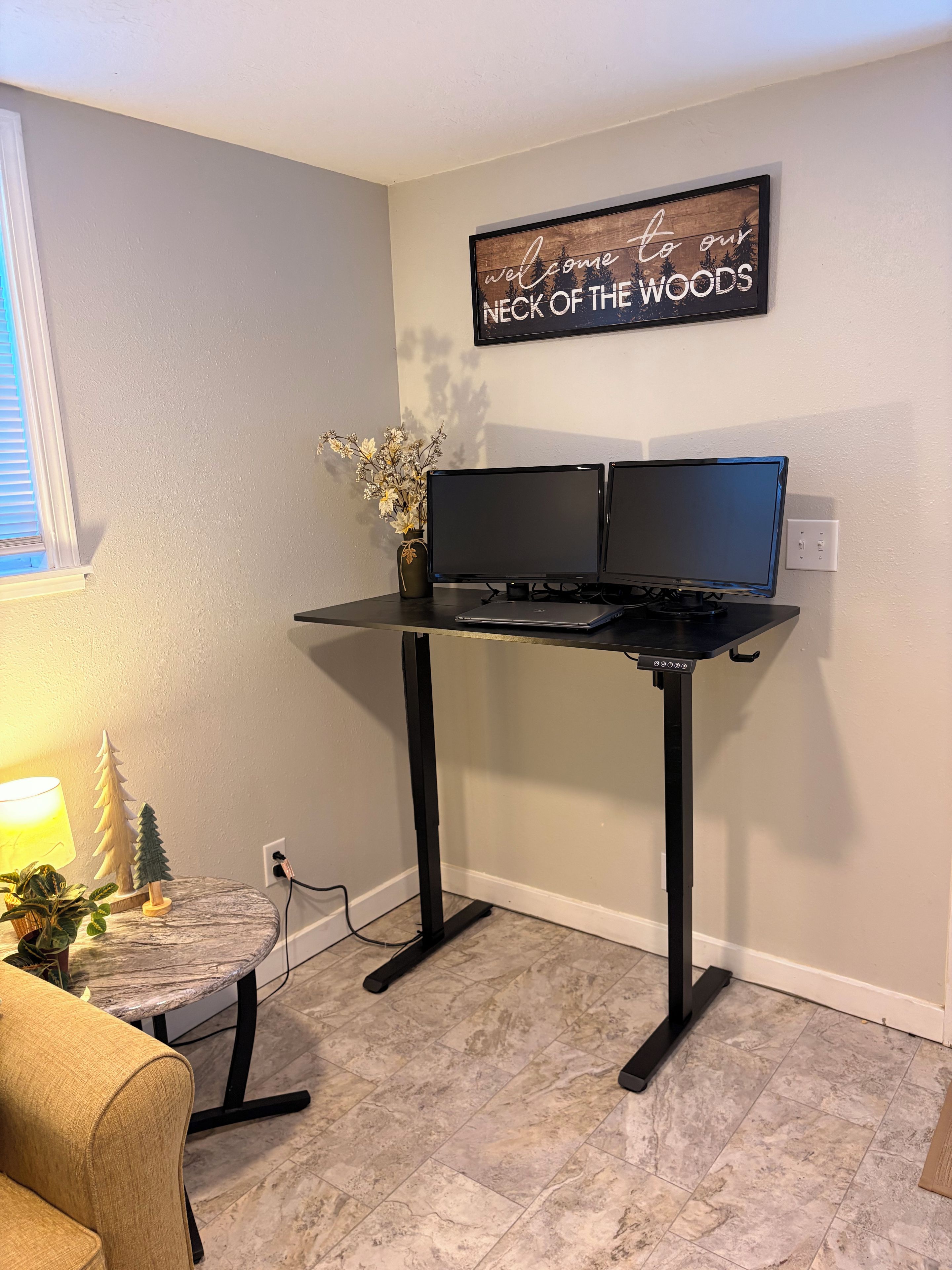 Un espacio de trabajo en casa con un escritorio, dos monitores, una computadora portátil, una planta pequeña y un letrero decorativo en la pared.