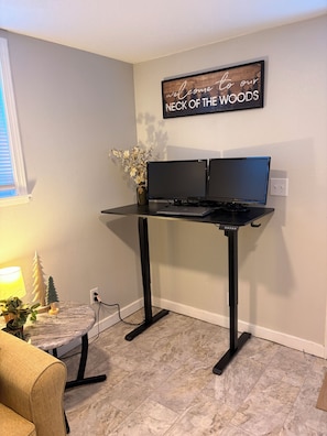 Un espacio de trabajo en casa con un escritorio, dos monitores, una computadora portátil, una planta pequeña y un letrero decorativo en la pared.