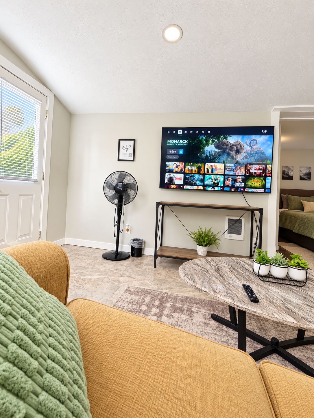 Una sala de estar con un televisor de pantalla plana montado en la pared, un ventilador de pie, una mesita con plantas en macetas y un sofá.