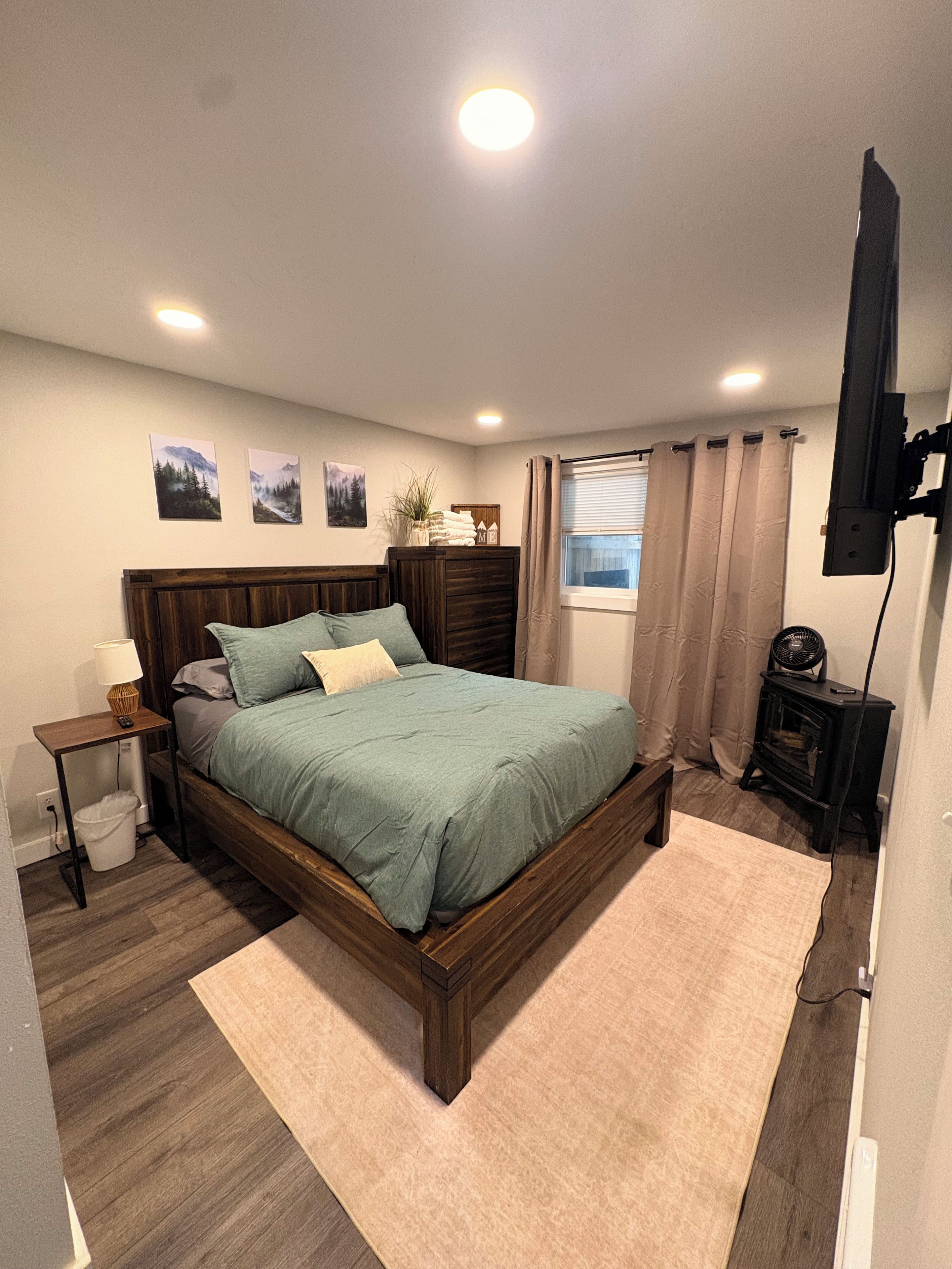 A bedroom with a wooden bed, a bedside table with a lamp, a wall-mounted TV, and a window with curtains.