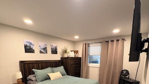 Ein Schlafzimmer mit einem hölzernen Bett, einem Nachttisch, einer Lampe, einem an der Wand montierten Fernseher und einem Fenster mit Vorhängen.
