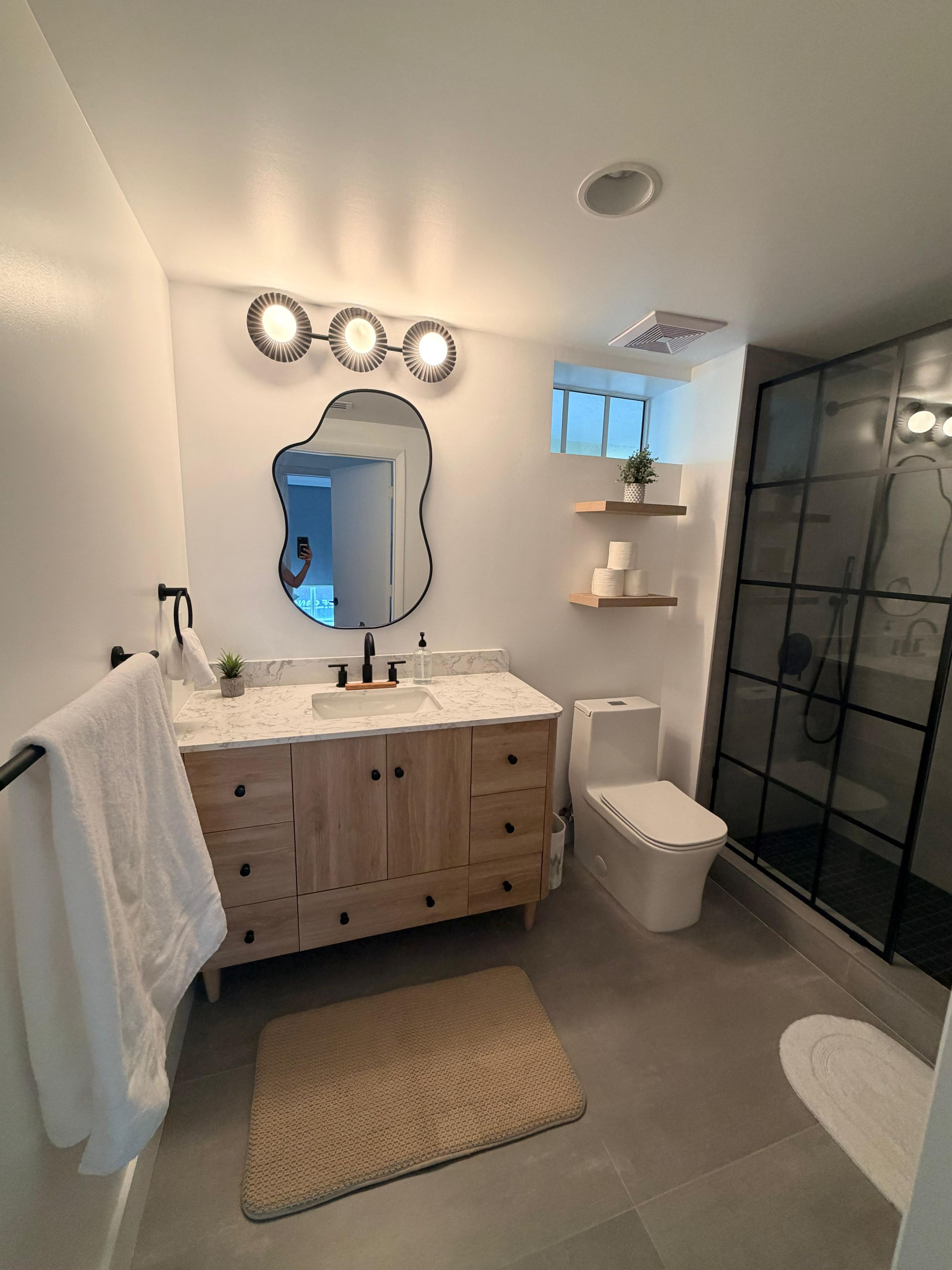 Une salle de bain moderne comprenant un meuble-lavabo, des toilettes, un espace douche et un grand miroir avec des lumières au-dessus.