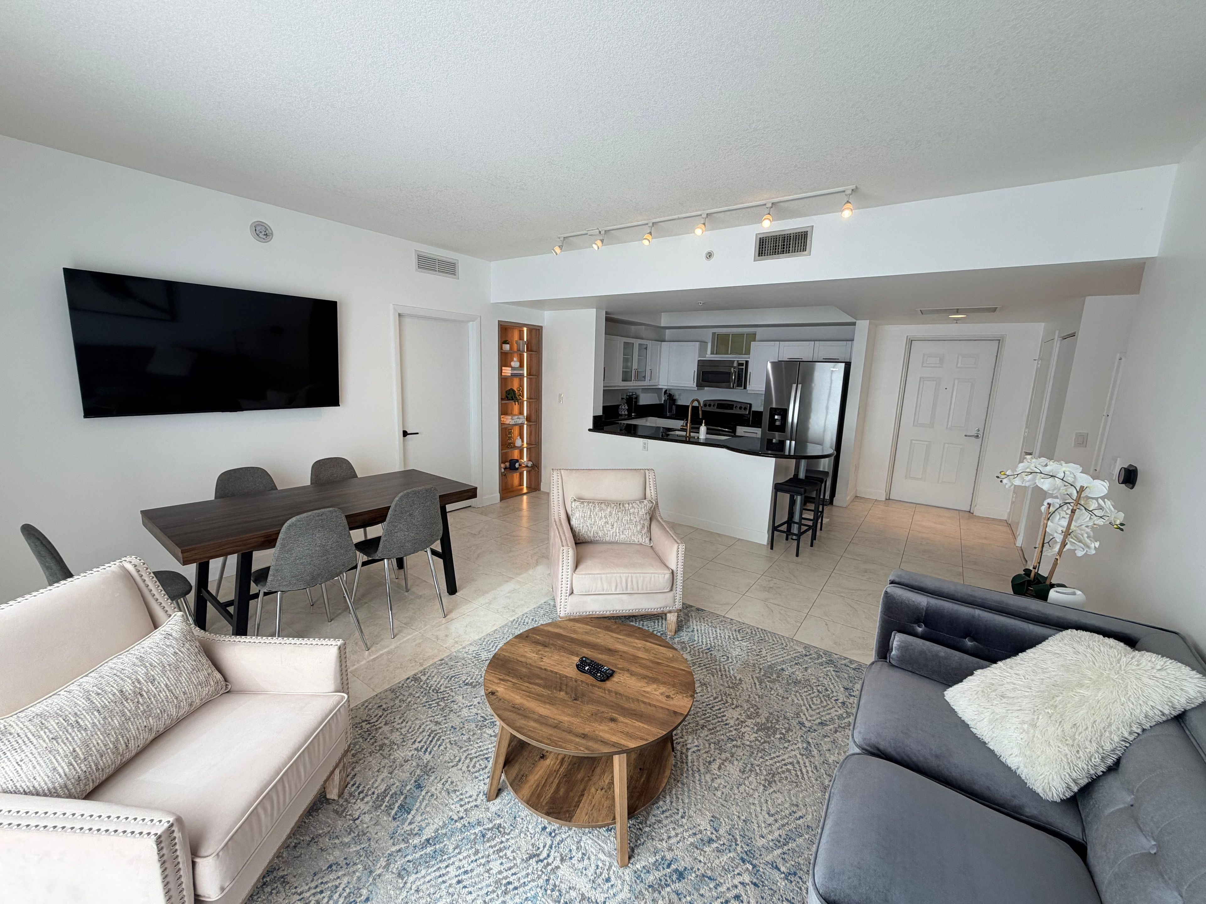 A modern living room with a dining area, kitchen, and a flat-screen TV mounted on the wall.