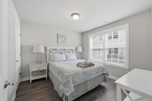 A bedroom with a bed, two bedside tables, a nightstand, a window with blinds, and a door. - VIBE 3BR | 2 Blks to Beach & 1 blck Atlantic Park (Virginia Beach)