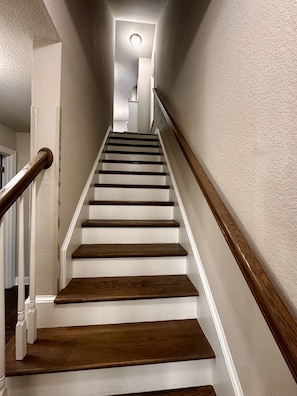 Una escalera con peldaños de madera y paredes blancas, que lleva a una plataforma con una lámpara.