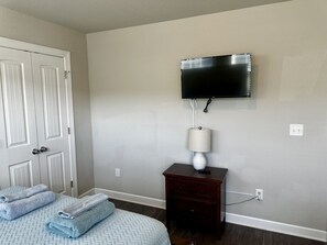 Une chambre à coucher avec un lit, un téléviseur fixé au mur, une table de chevet avec une lampe et une porte sur laquelle sont accrochées des serviettes.