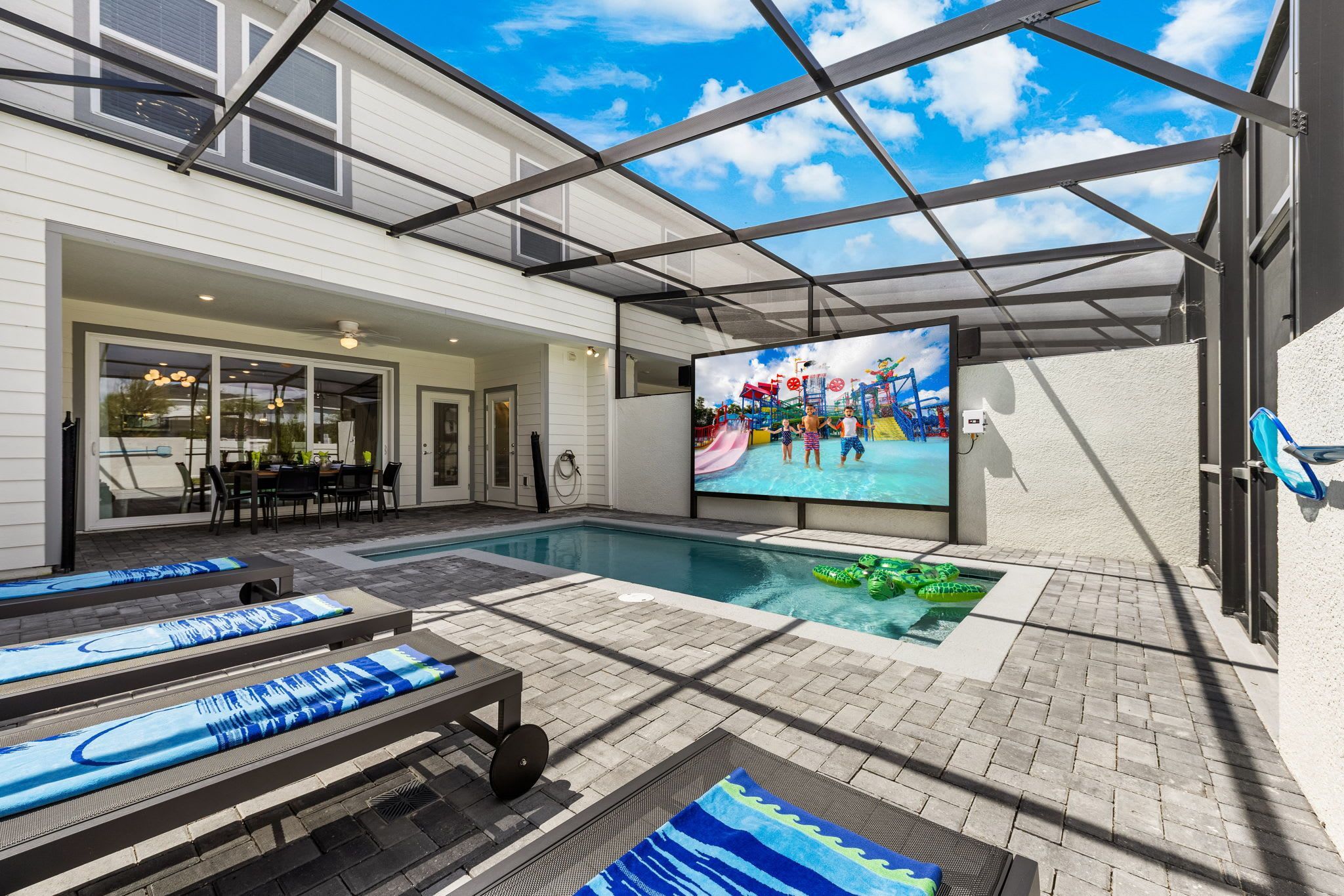 A pool area with a large screen displaying a water park, lounge chairs, and a dining area.