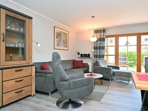 A modern living room with a grey sofa, a wooden cabinet with glass doors, a dining table, and a large window with curtains. - 4.3/t2 Holiday Park "freesenbruch" (Zingst)