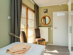 A hotel room with a bed, a chair, a radiator, a door, and a window with curtains. - 4.3/t2 Holiday Park "freesenbruch" (Zingst)