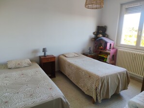 Une chambre avec deux lits simples, une table de chevet avec une lampe, une commode et une fenêtre avec des stores.
