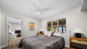 Une chambre à coucher comprenant un lit, un ventilateur de plafond, une fenêtre avec des stores, une table de chevet avec une lampe et une œuvre d’art accrochée au mur.