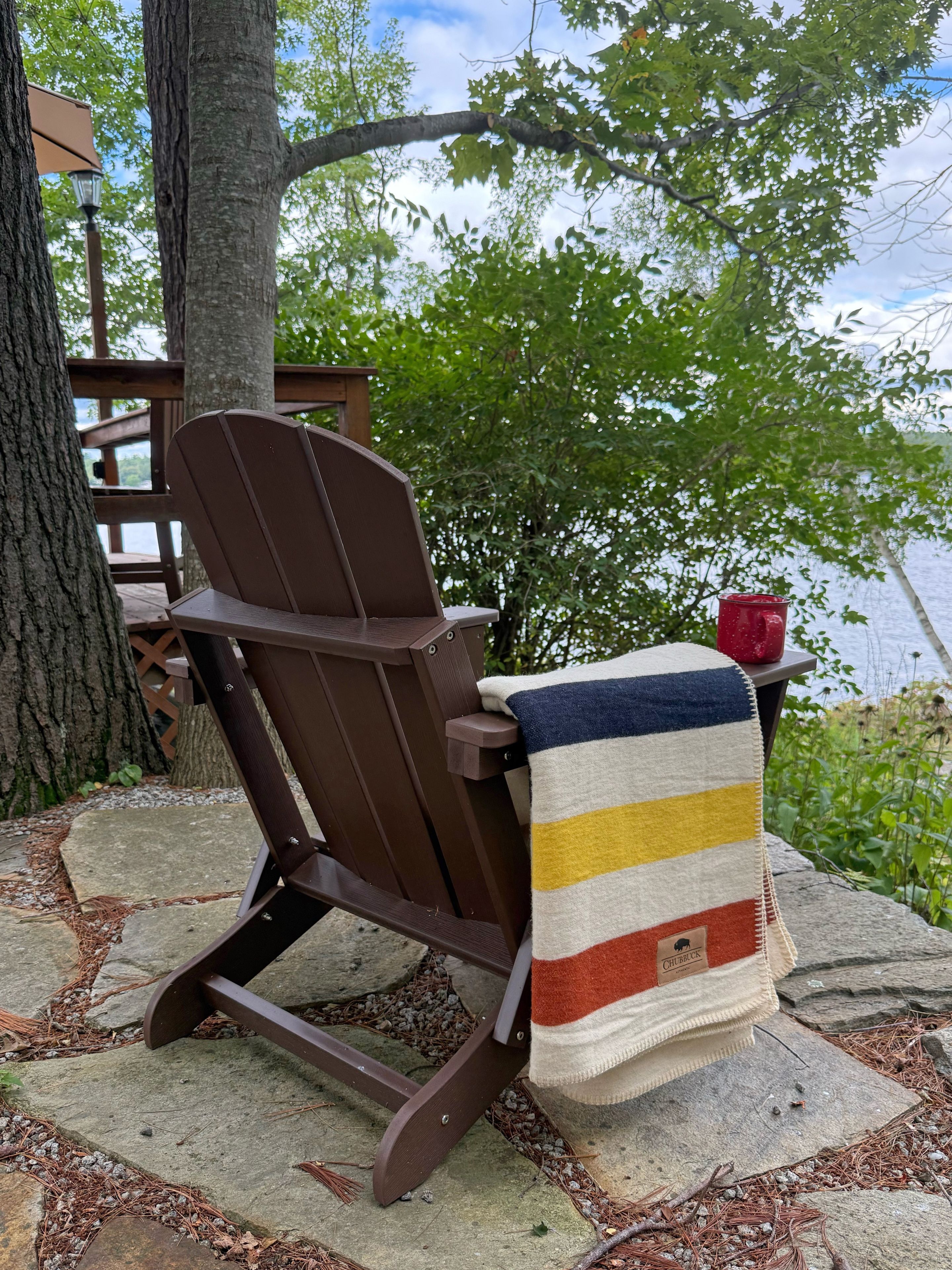 A wooden Adirondack chair with a striped blanket and a red mug on a stone ledge.