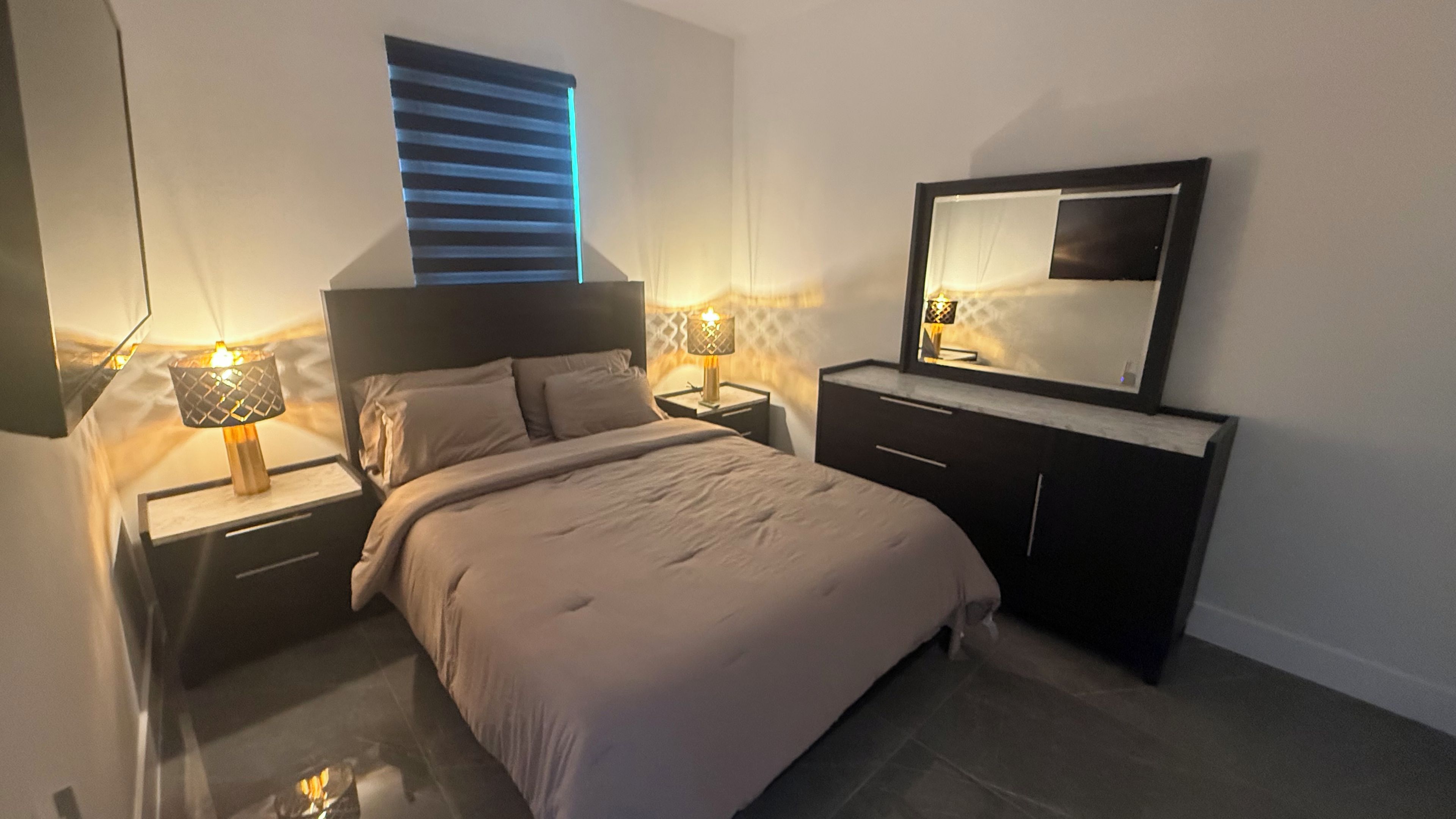 Un dormitorio con una cama, mesitas de noche, un aparador, un espejo y una ventana con persianas.