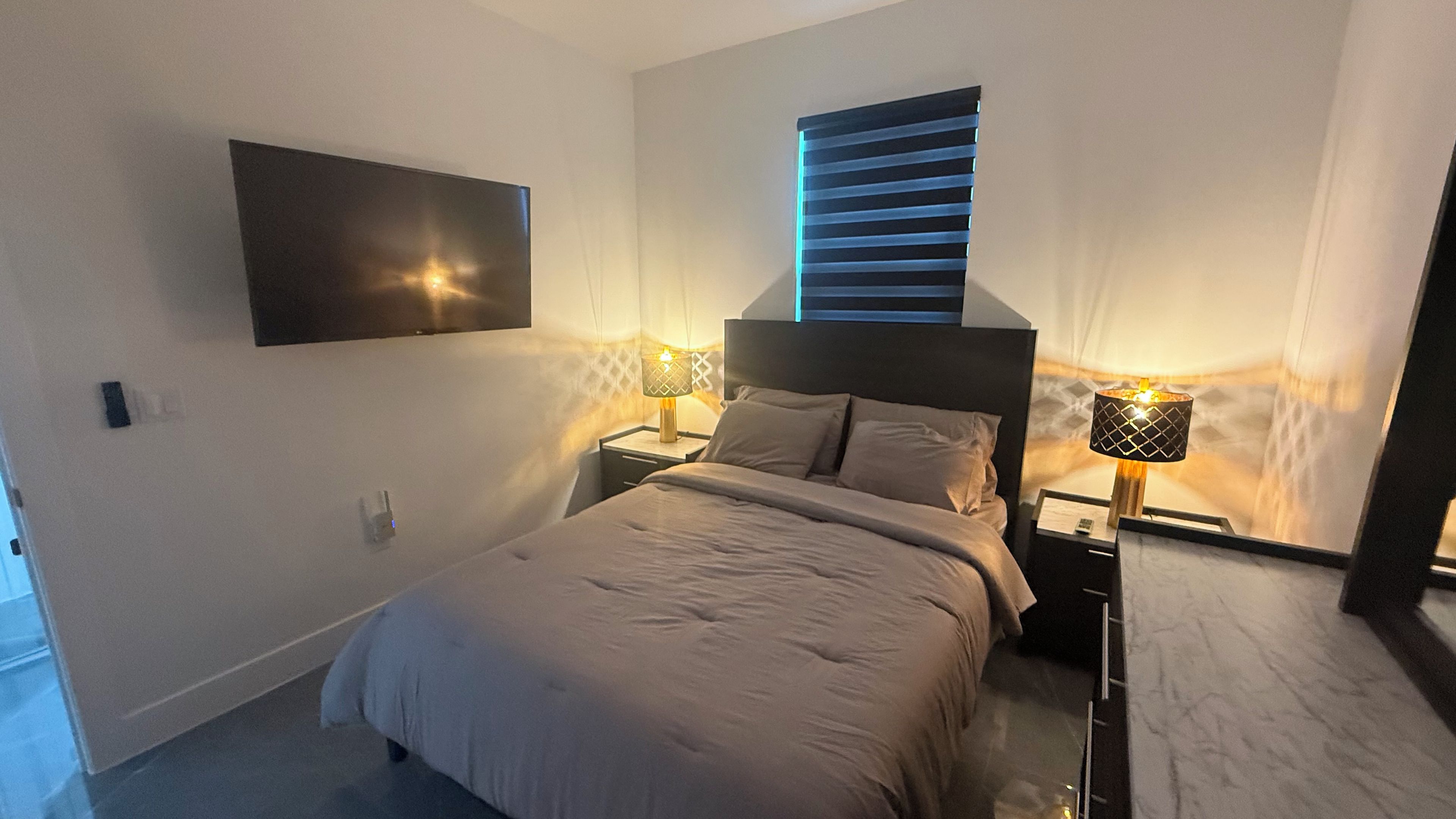A bedroom with a bed, two bedside tables, a wall-mounted TV, and a window with blinds.