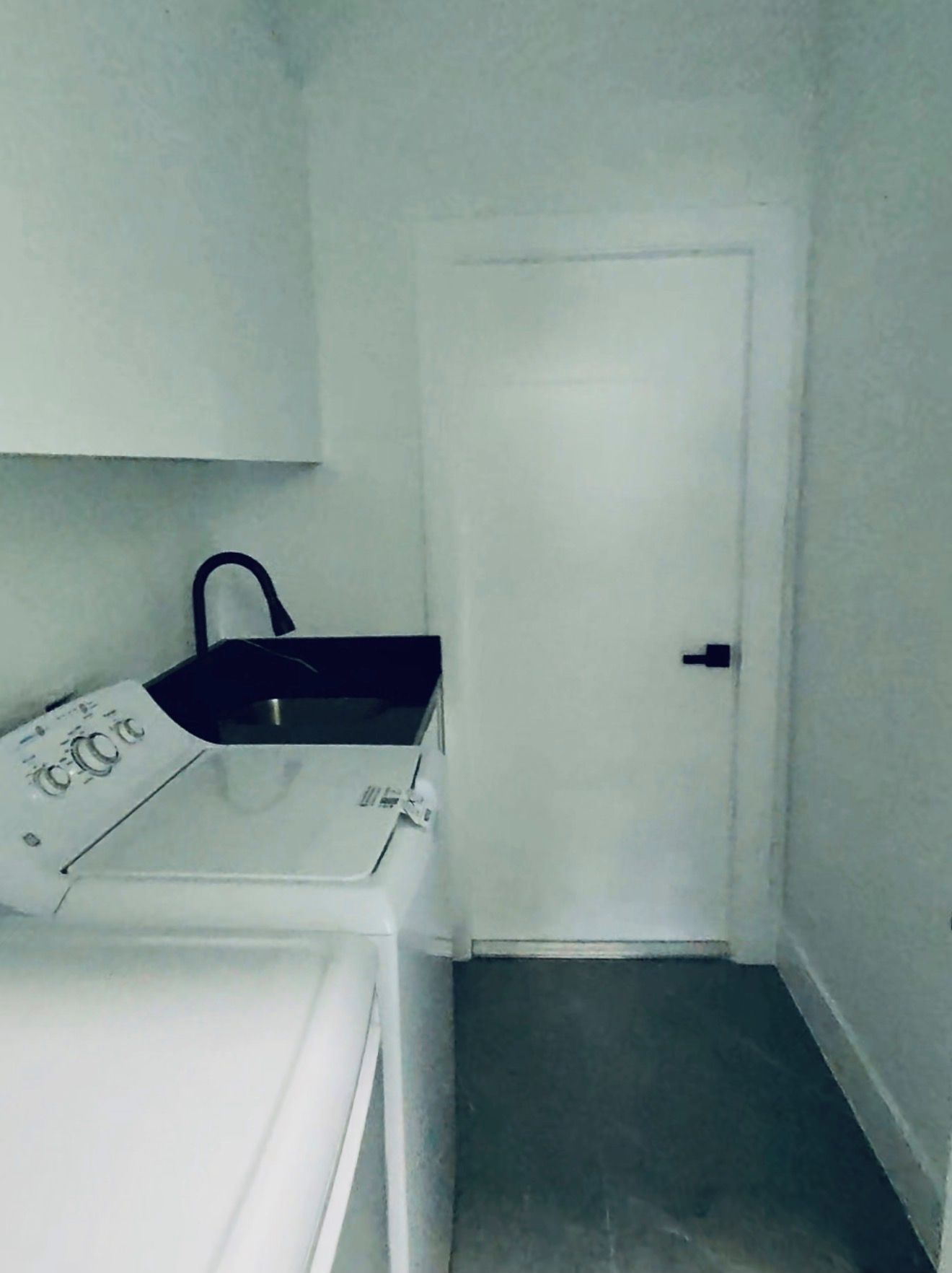 A small, narrow room with a white door, a white washing machine, and a white cabinet with a black handle.