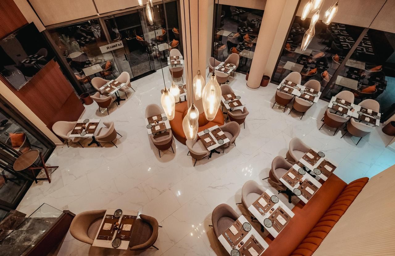 A spacious dining area with multiple tables and chairs, a central light fixture, and a view of the restaurant through glass walls.