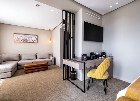 A modern hotel room with a sofa, desk, chair, and a flat-screen TV mounted on the wall.