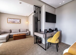 A modern hotel room with a sofa, desk, chair, and a flat-screen TV mounted on the wall.