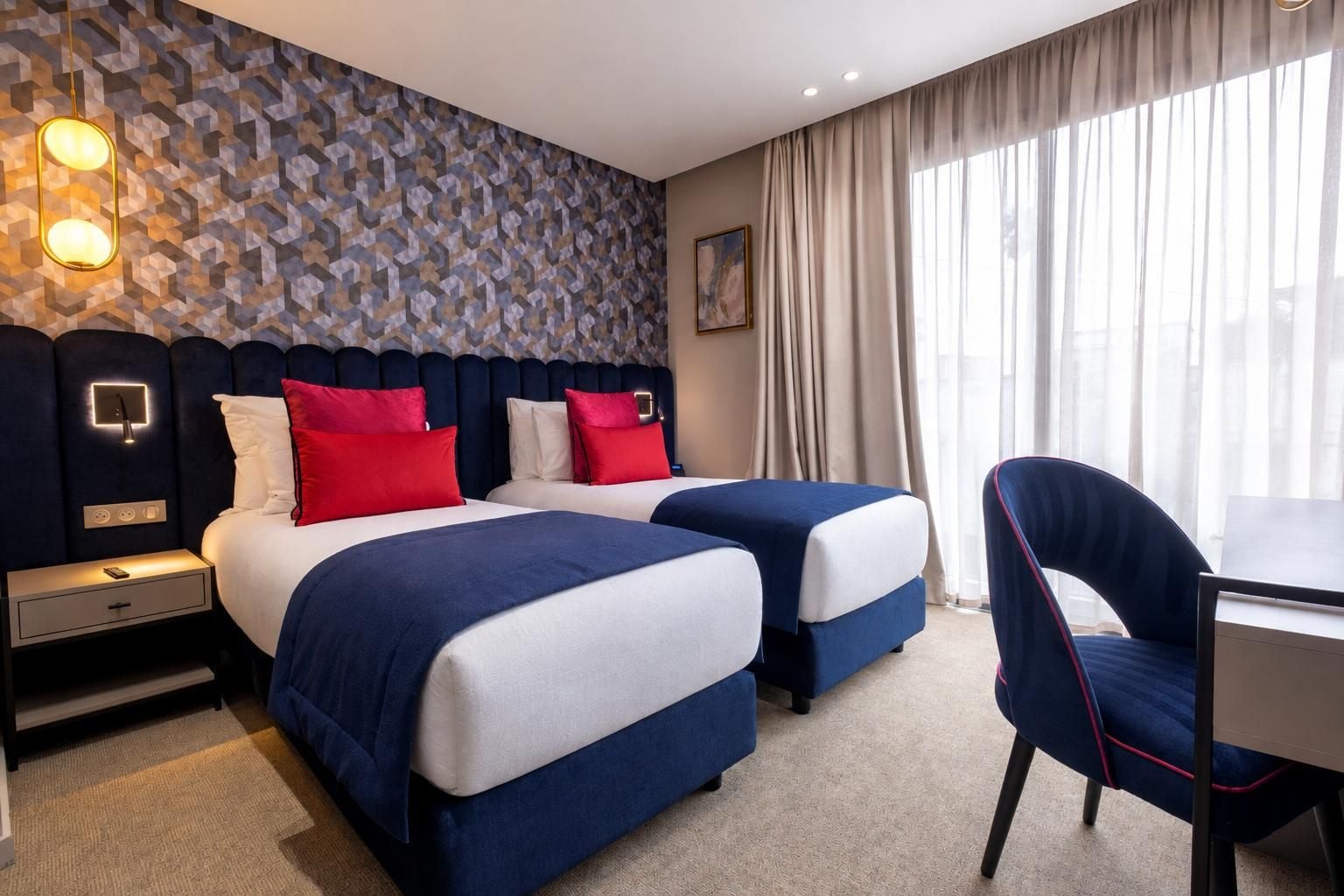 A hotel room with two beds, a desk, a chair, a patterned wall, and a window with curtains.