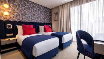 A hotel room with two beds, a desk, a chair, a patterned wall, and a window with curtains.