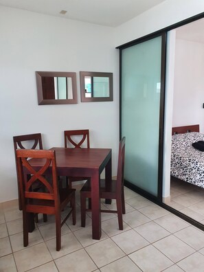 Zona pranzo con tavolo e sedie in legno, due specchi fissati alla parete e una porta vetrata scorrevole che dà su una camera da letto.