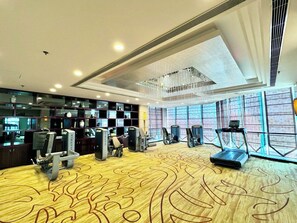 Academia de hotel com equipamentos de ginástica, carpete estampado e amplas janelas.