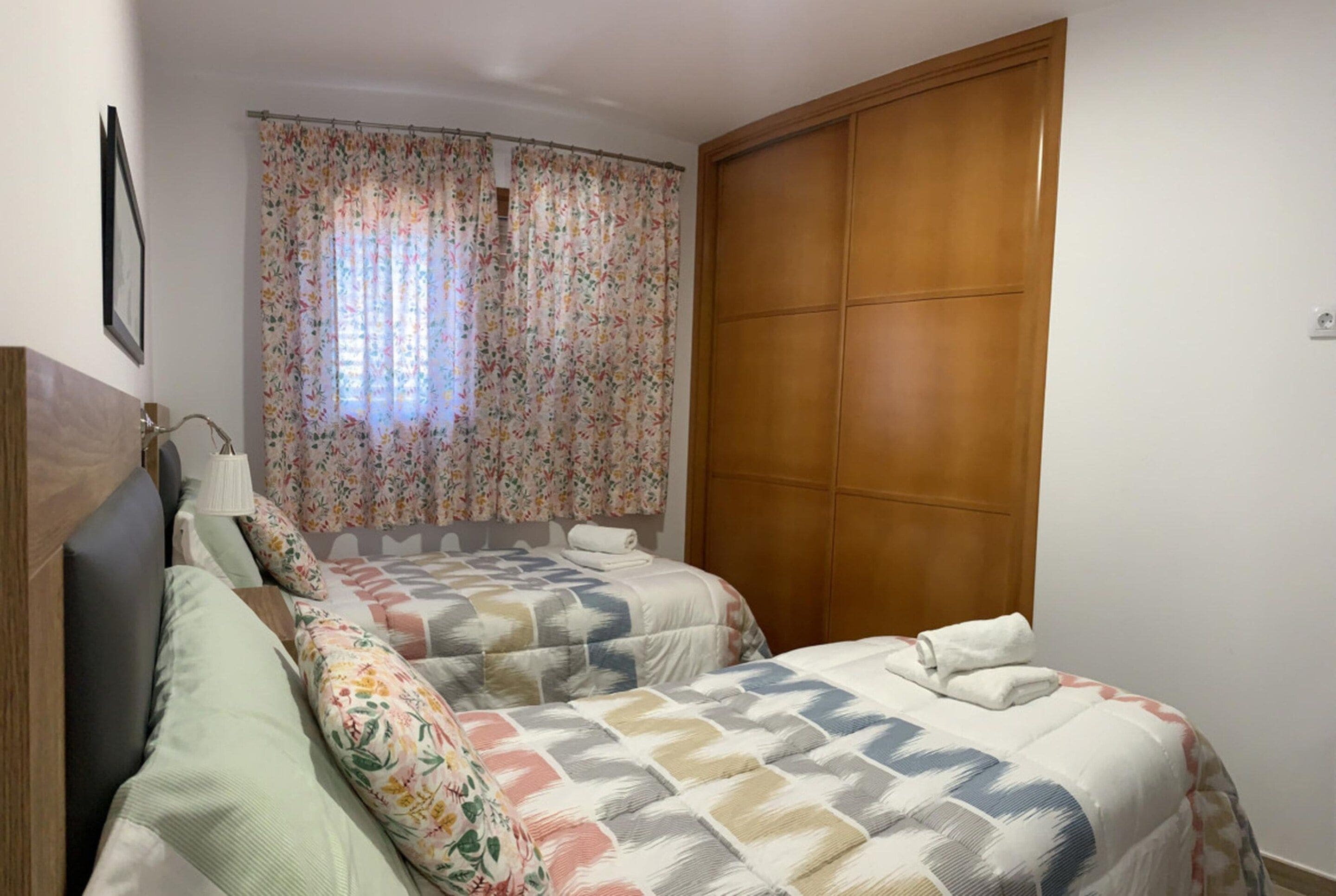 Une chambre avec deux lits, une armoire en bois et une fenêtre ornée de rideaux à motifs floraux.