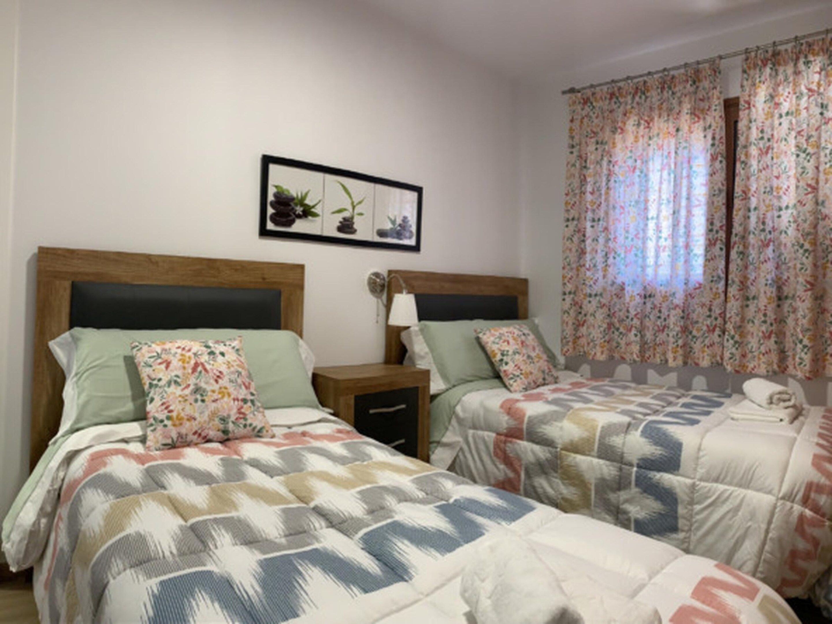 Ein Doppelzimmer mit zwei Betten, jedes mit einem floralen Kopfkissen und einer gemusterten Decke. Es gibt einen Nachttisch mit einer Lampe sowie ein gerahmtes Bild von Pflanzen an der Wand.