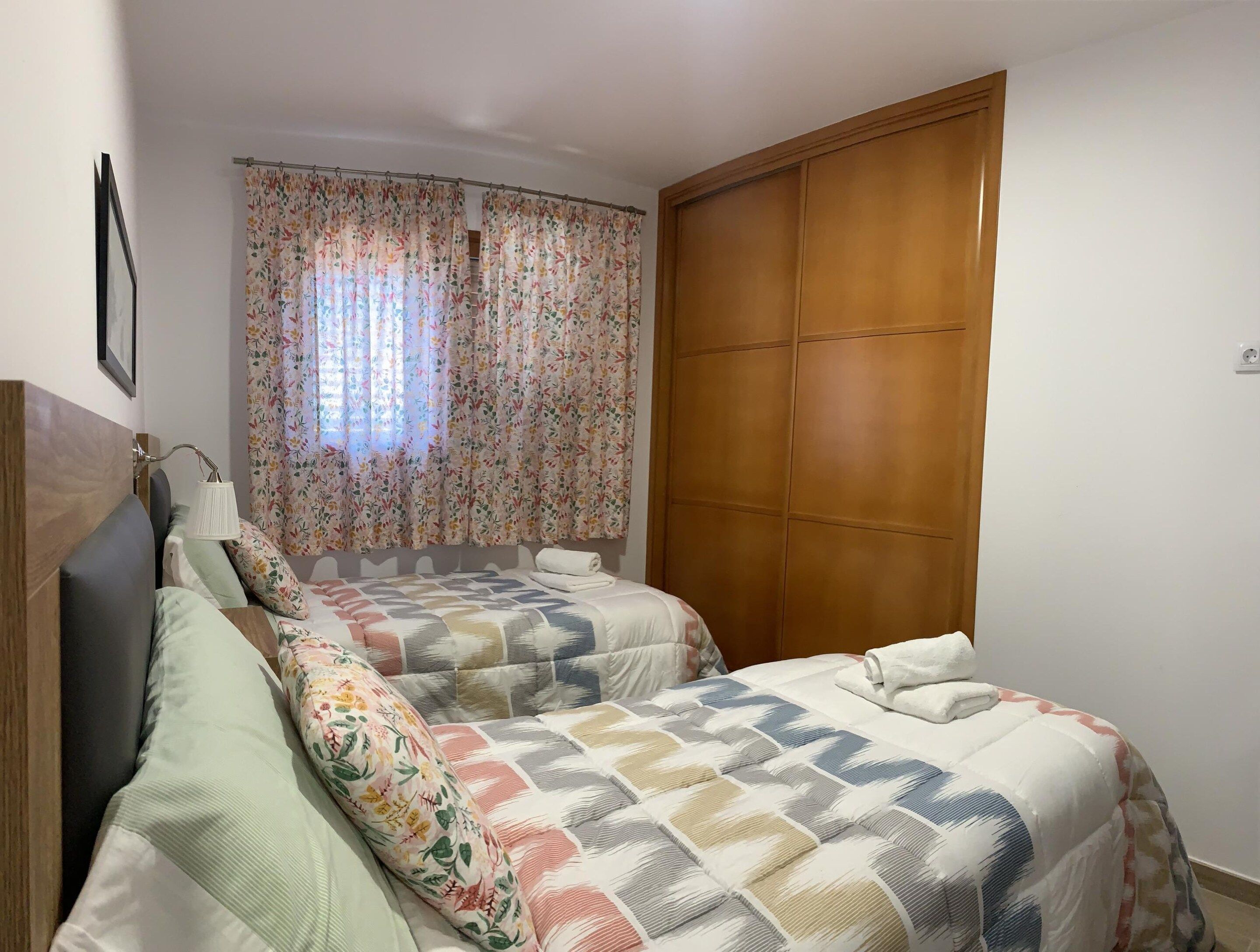 Un dormitorio con dos camas, un armario de madera, un ventanal con cortinas florales y una lámpara de pared.