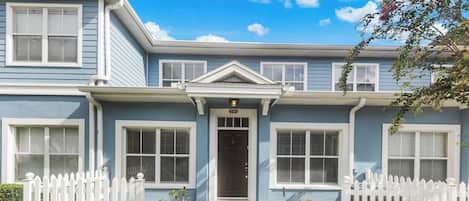 A two-story residential building with a front porch, white picket fence, and a number '27-104' above the door.