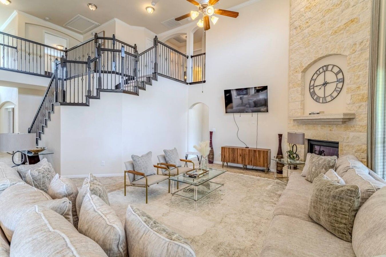 A spacious living room with a high ceiling, a staircase, a flat-screen TV, a fireplace, and a stone wall.