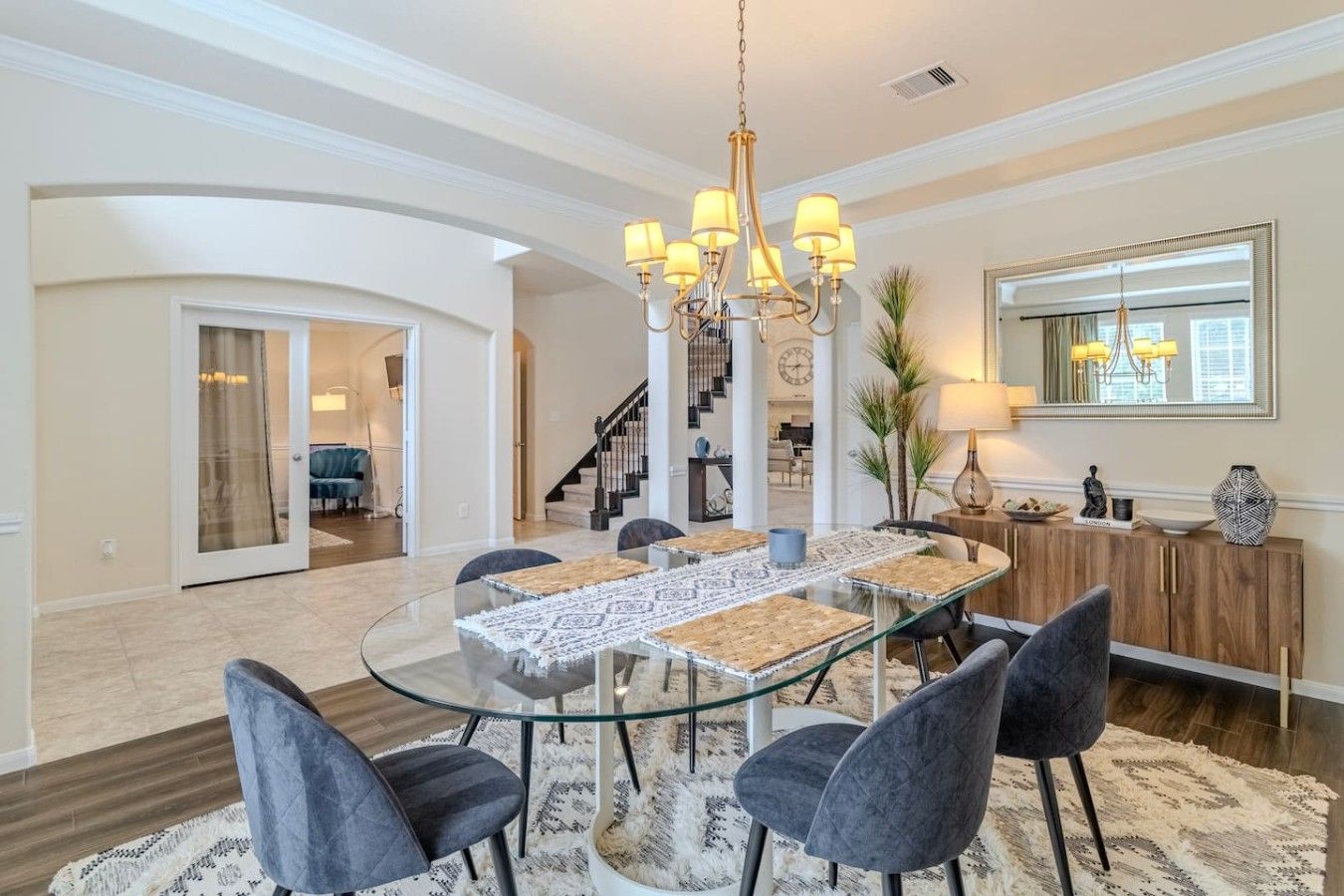 A dining area with a glass table, six chairs, a chandelier, and a large mirror on the wall.