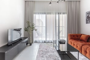 Een moderne woonkamer met een flatscreen televisie, een bank, een glazen salontafel en een groot raam met dunne gordijnen.
