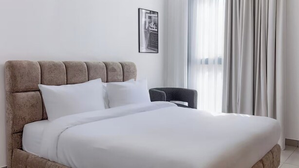 A neatly made bed with a headboard, a single chair, and a window with curtains. - Sahab - Sunset 1BR at Al mamsha Sharjah (Sharjah)