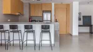 A modern kitchen with wooden cabinets, a stainless steel refrigerator, and a bar area with four stools. - Sahab - Sunset 1BR at Al mamsha Sharjah (Sharjah)