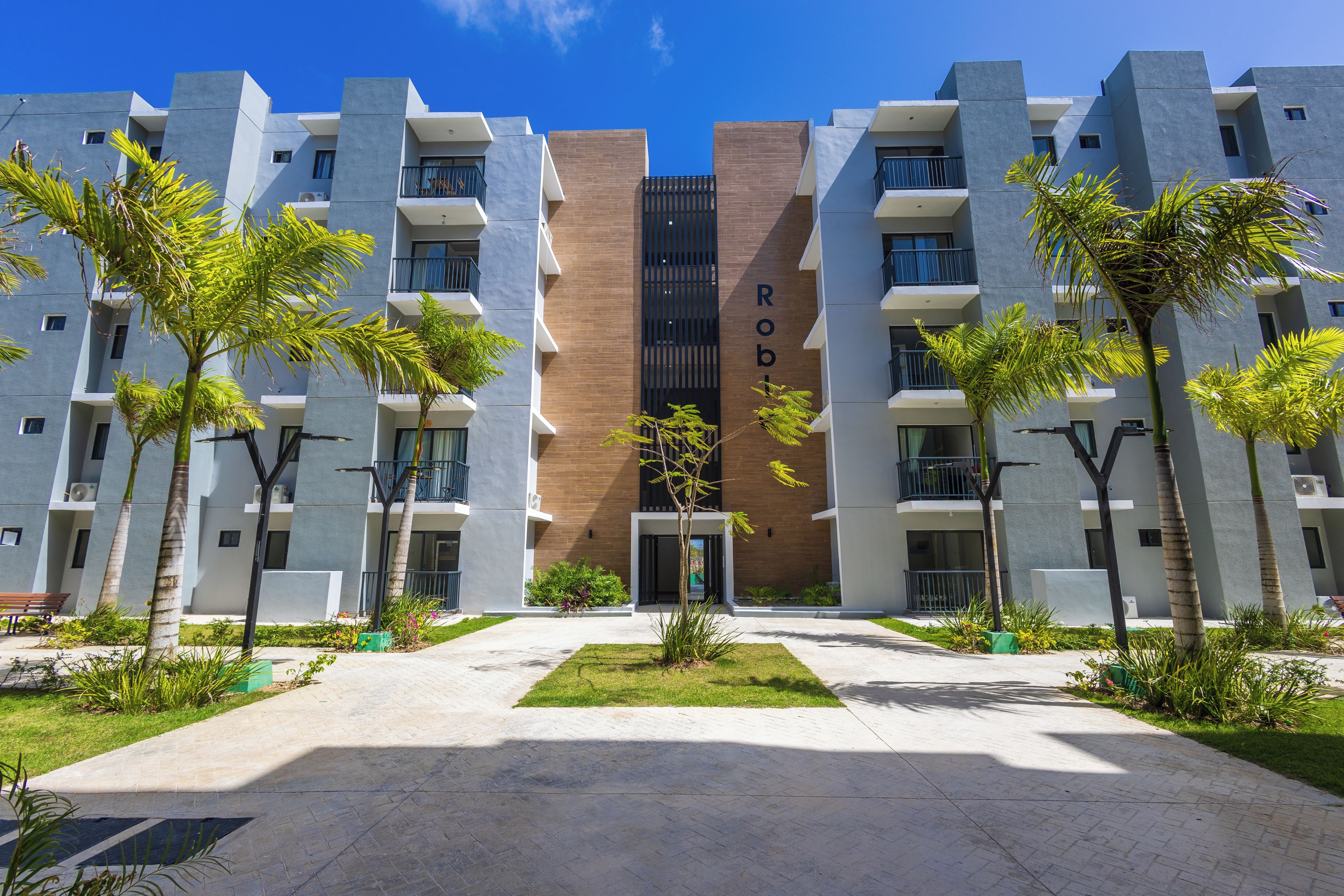 A modern multi-story building with a central entrance, surrounded by a landscaped area with palm trees and a paved walkway.